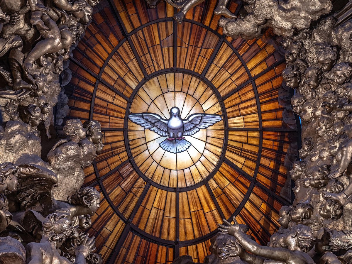 A stained-glass window of the Holy Spirit as a white dove descending with outstretched wings, in a glowing amber oval framed by gilded sculpted angels and clouds