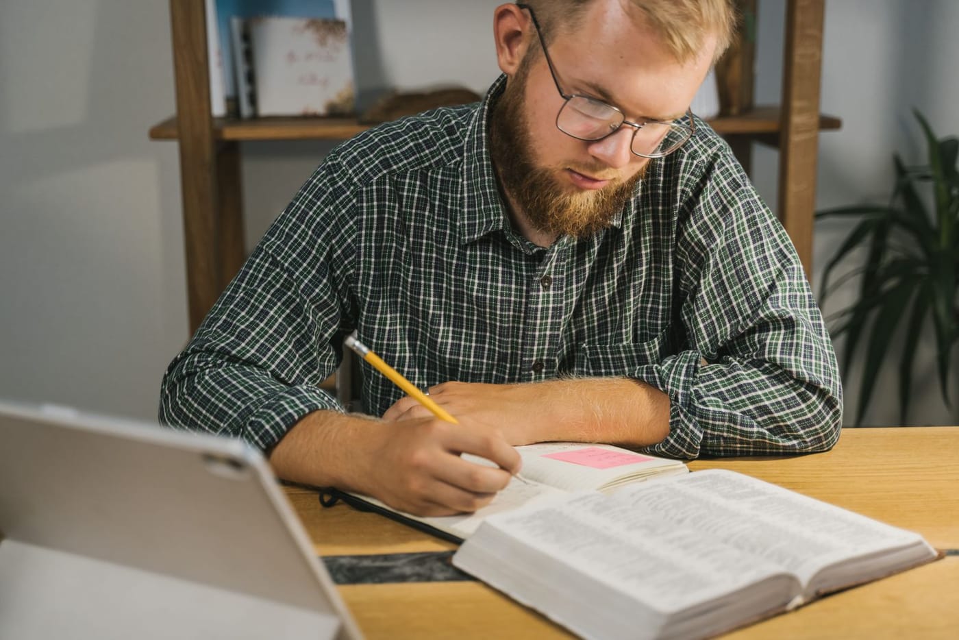 A man in glasses at a desk, pencil in hand, writing notes beside a large open Bible